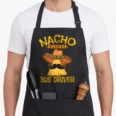 Nacho chips lover apron - Cartoon nacho chip in sombrero and mustache with Nacho Average Bus Driver text in yellow — shown on a black cotton canvas apron against a white background