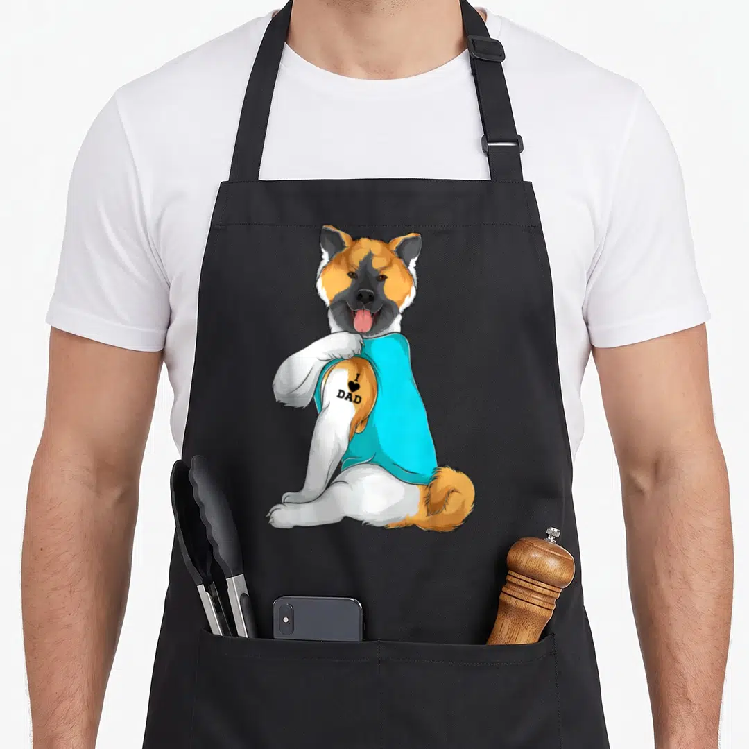 Akita tattoo dog apron - Cartoon Akita lifting cyan shirt to reveal I Love Dad tattoo on shoulder — shown on a black cotton canvas apron against a white background