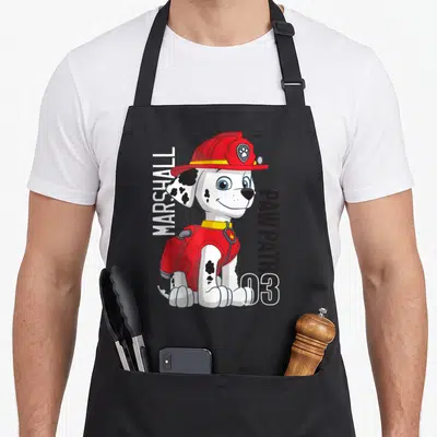 Character marshalls apron - Cartoon dalmatian in red gear with vertical MARSHALL PAW PATROL 03 text — shown on a black cotton canvas apron against a white background
