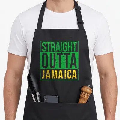 Straight creole apron - Stacked STRAIGHT OUTTA JAMAICA text in green grey and yellow distressed font — shown on a black cotton canvas apron against a white background
