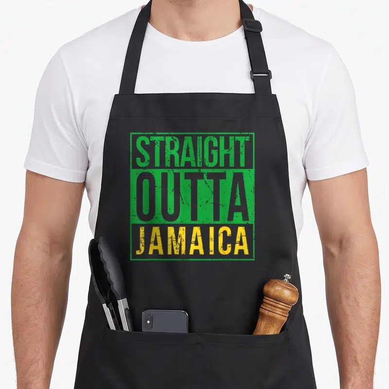 Straight creole apron - Stacked STRAIGHT OUTTA JAMAICA text in green grey and yellow distressed font — shown on a black cotton canvas apron against a white background