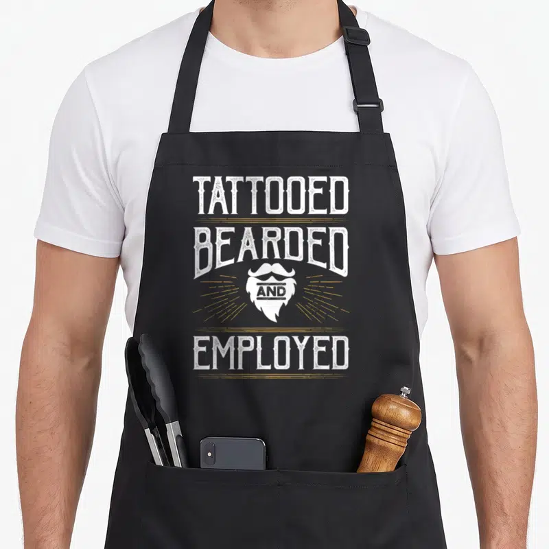 Tattoo beard apron - Stacked white distressed text reading Tattooed Bearded and Employed with a beard graphic — shown on a black cotton canvas apron against a white background
