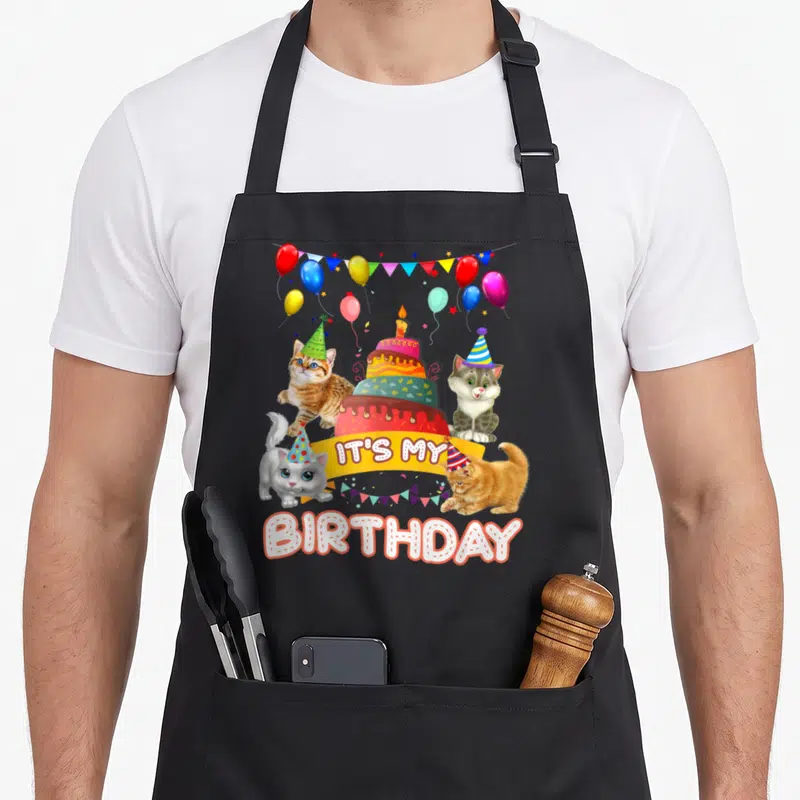 Birthday kitten apron - Four cats in party hats surrounding a cake with IT'S MY BIRTHDAY text — shown on a black cotton canvas apron against a white background