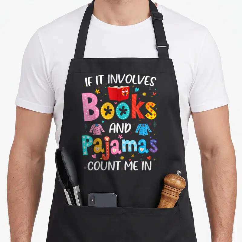 Involve pajama day apron - Stacked text reading If It Involves Books And Pajamas Count Me In with cartoon book — shown on a black cotton canvas apron against a white background