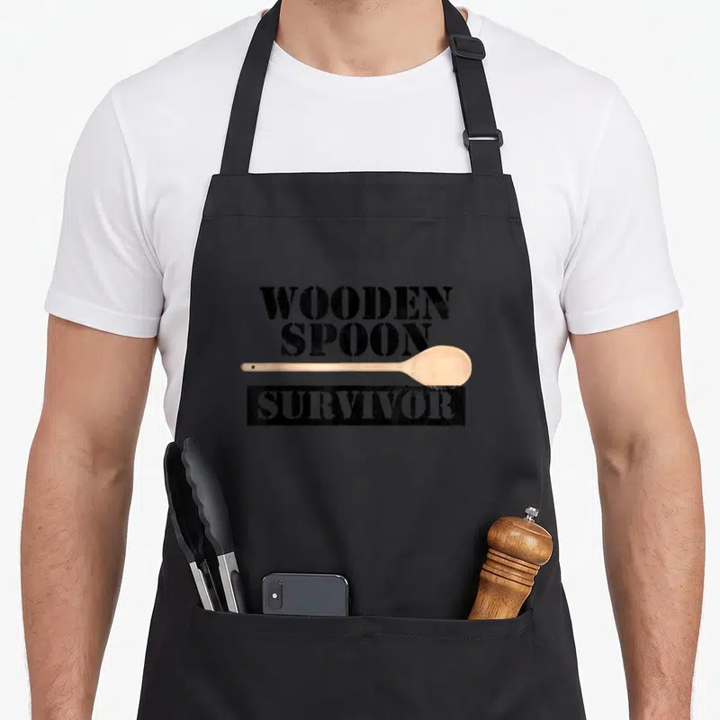 Filipino wooden apron - WOODEN SPOON SURVIVOR text with horizontal light brown spoon and distressed black block — shown on a black cotton canvas apron against a white background