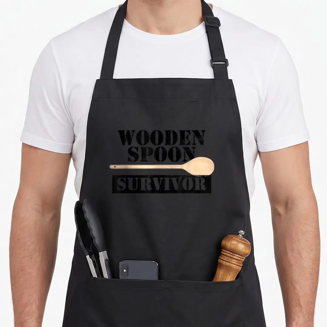 Filipino wooden apron - WOODEN SPOON SURVIVOR text with horizontal light brown spoon and distressed black block — shown on a black cotton canvas apron against a white background