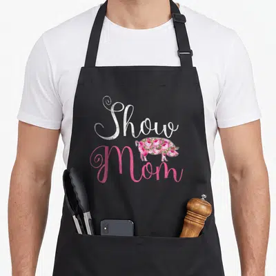 Pig cooking mama apron - Show Mom text with pink and white floral pig filling the silhouette — shown on a black cotton canvas apron against a white background