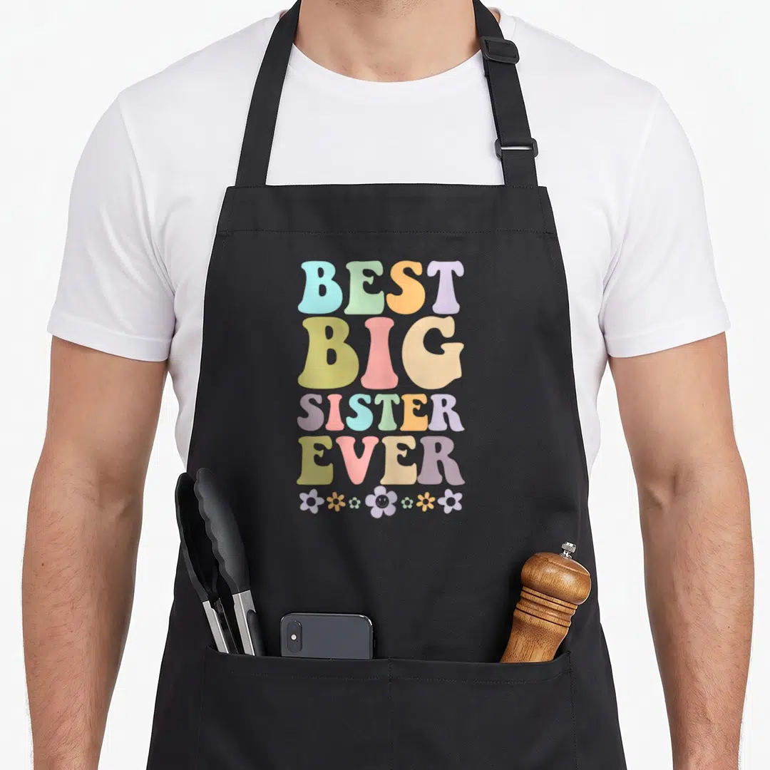 Announcement family aprons - Stacked BEST BIG SISTER EVER text in pastel colors above seven small flowers — shown on a black cotton canvas apron against a white background