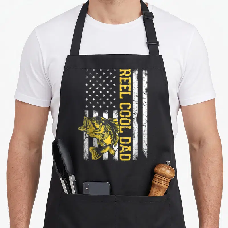 Reel fishing father day apron - Yellow fish and white stripes with vertical REEL COOL DAD text in distressed style — shown on a black cotton canvas apron against a white background
