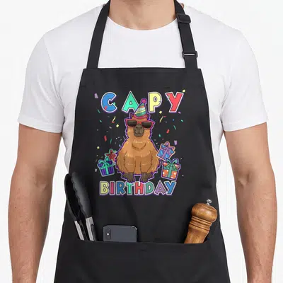 Capybara capy birthday apron - Cartoon capybara in sunglasses and party hat with CAPY BIRTHDAY text and gift boxes — shown on a black cotton canvas apron against a white background
