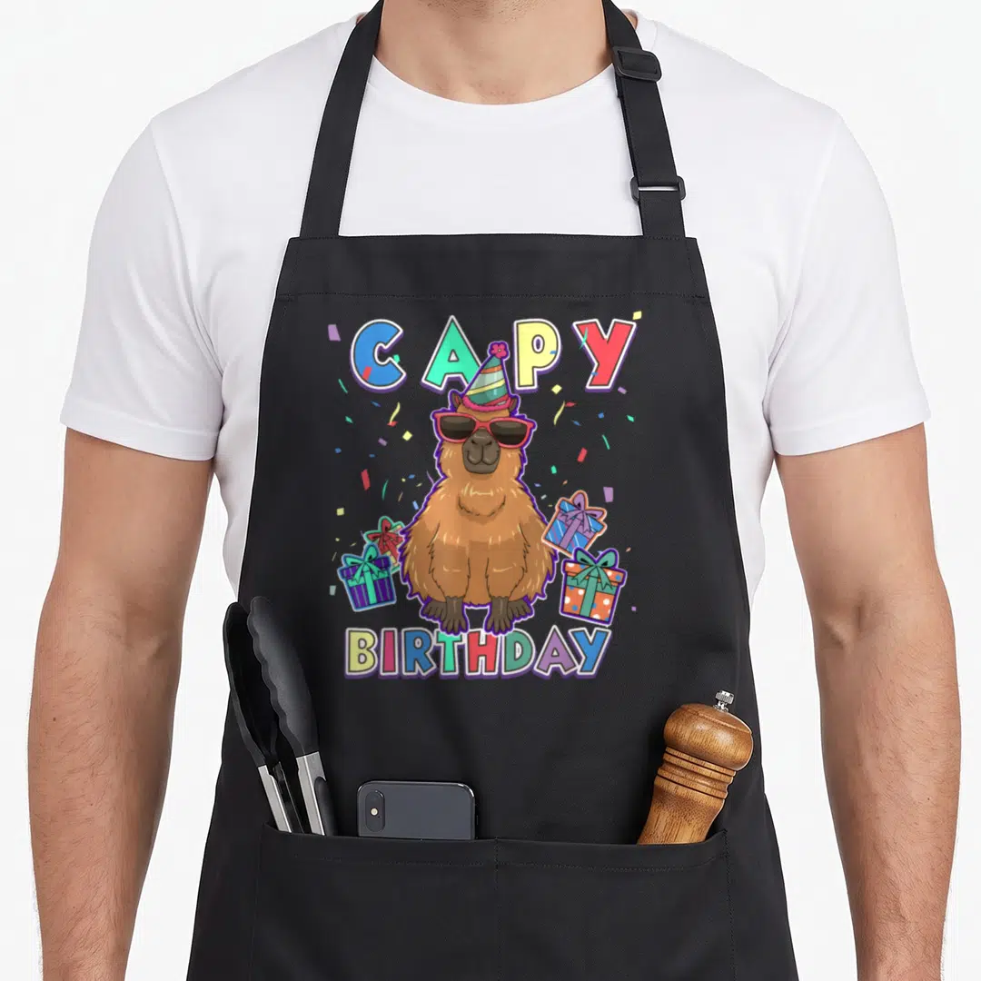 Capybara capy birthday apron - Cartoon capybara in sunglasses and party hat with CAPY BIRTHDAY text and gift boxes — shown on a black cotton canvas apron against a white background