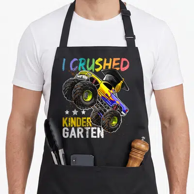 Crushed truck aprons - Monster truck in grad cap with rainbow I CRUSHED text and mud splatters — shown on a black cotton canvas apron against a white background