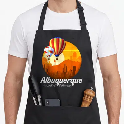 Festival mexico apron - Two striped balloons over orange desert landscape with white stacked text reading Albuquerque — shown on a black cotton canvas apron against a white background