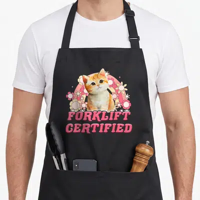 Forklift cat apron - Cartoon orange cat with pink rainbow, white flowers, and FORKLIFT CERTIFIED text — shown on a black cotton canvas apron against a white background