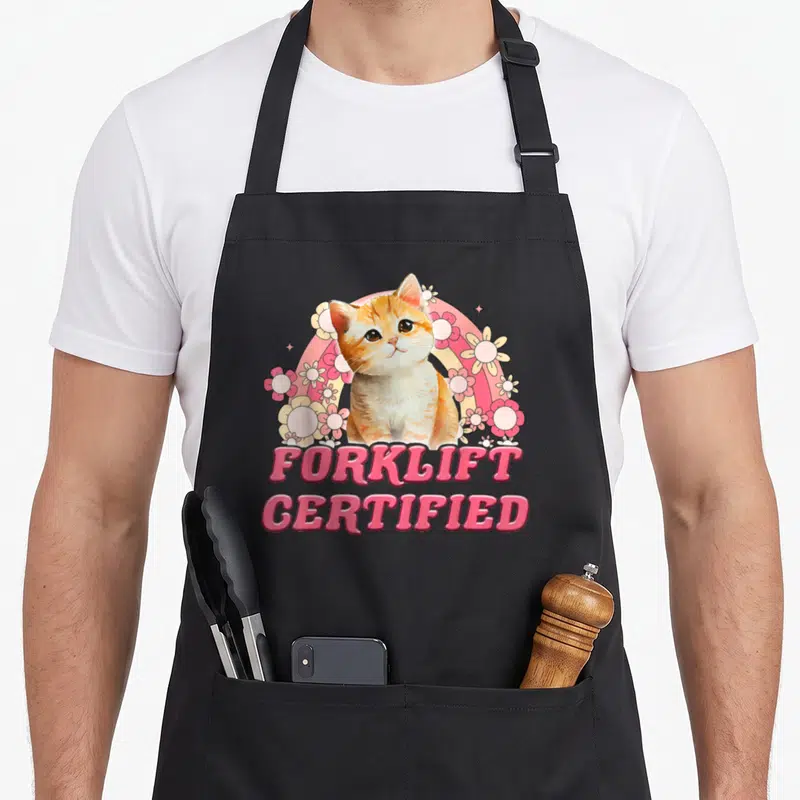 Forklift cat apron - Cartoon orange cat with pink rainbow, white flowers, and FORKLIFT CERTIFIED text — shown on a black cotton canvas apron against a white background