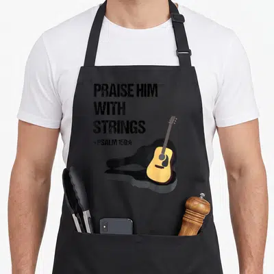 Guitar player christian apron - Stacked Praise Him With Strings text beside yellow acoustic guitar leaning on black case — shown on a black cotton canvas apron against a white background