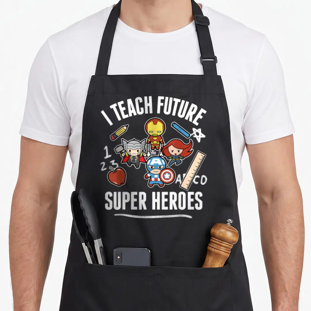 Teach heroe apron - I TEACH FUTURE SUPER HEROES text with chibi characters, pencils, ruler, and apple — shown on a black cotton canvas apron against a white background