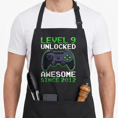 Level unlocked video birthday apron - Game controller centered between LEVEL 9 UNLOCKED and AWESOME SINCE 2012 text — shown on a black cotton canvas apron against a white background