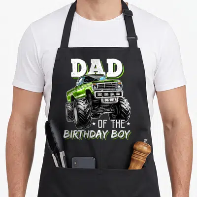 Cute birthday truck apron - Green monster truck illustration with Dad of the Birthday Boy text and stars — shown on a black cotton canvas apron against a white background