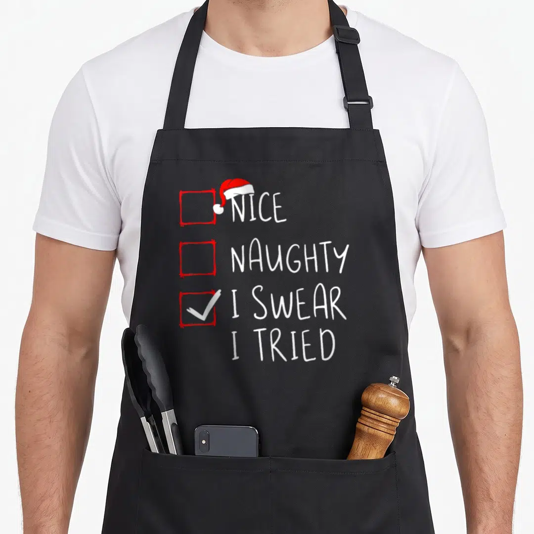 Swear santa claus apron - Red checkboxes with Santa hat and white text Nice Naughty I Swear I Tried — shown on a black cotton canvas apron against a white background