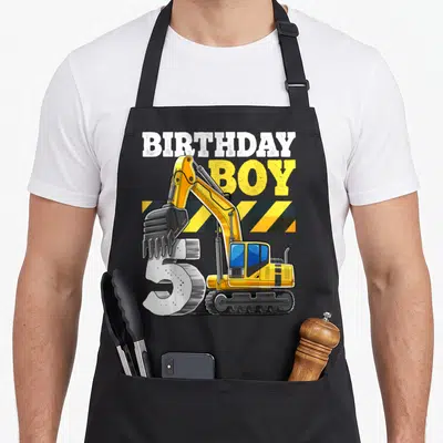 5th vehicle apron - Birthday Boy text over yellow excavator overlapping number 5 with caution stripes — shown on a black cotton canvas apron against a white background
