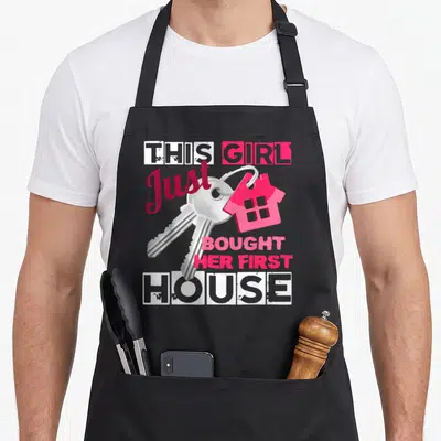 Bought novelty apron - Silver keys and pink house charm surrounded by stacked white and pink text on black fabric — shown on a black cotton canvas apron against a white background