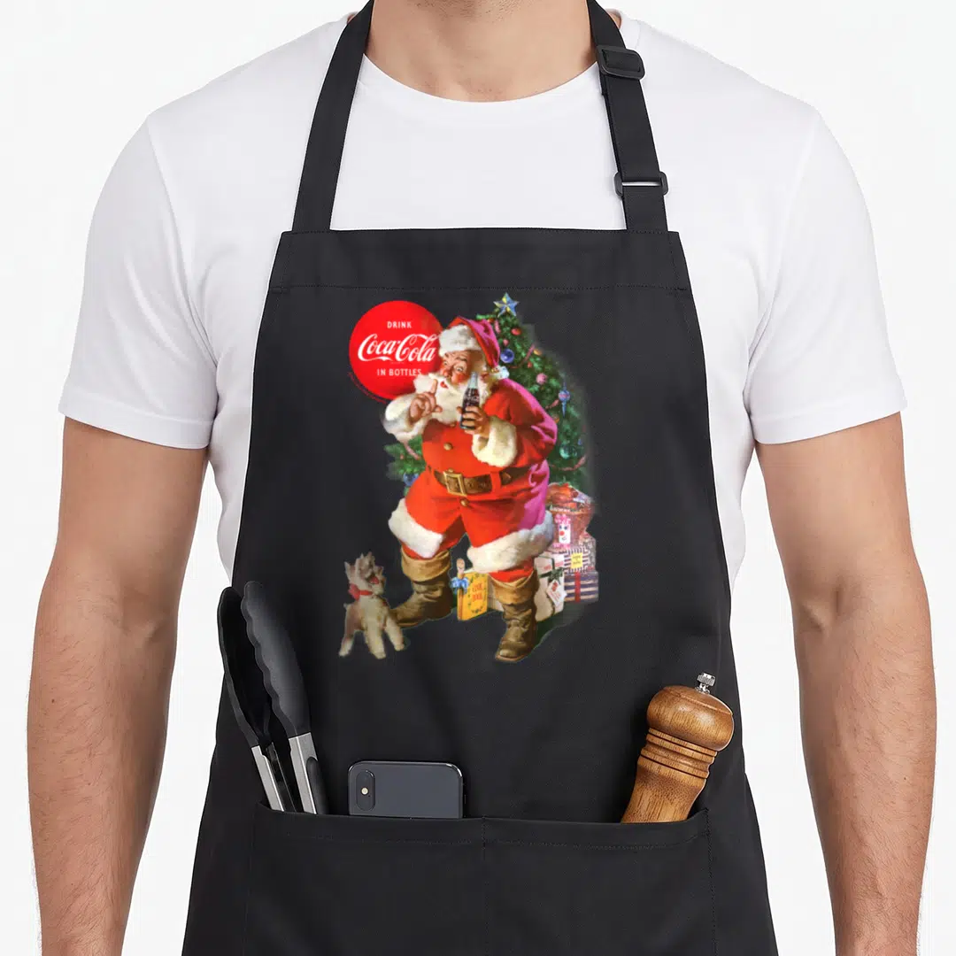 Coca santa claus apron - Santa holding bottle near dog cookbook and tree with red logo — shown on a black cotton canvas apron against a white background