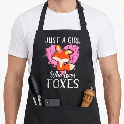 Funny fox print apron - Cartoon orange fox with flower crown in front of pink heart and white text — shown on a black cotton canvas apron against a white background