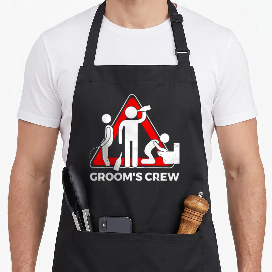 Groom crew apron - Red warning triangle with three stick figures and GROOM'S CREW text — shown on a black cotton canvas apron against a white background