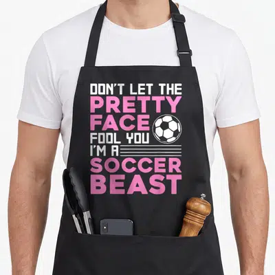 Sport soccer player accessories apron - Stacked pink and white text reading DON'T LET THE PRETTY FACE FOOL YOU I'M — shown on a black cotton canvas apron against a white background