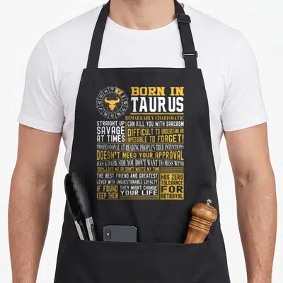 Fact taurus aprons - Zodiac bull wheel and Born in Taurus text in yellow and white block typography — shown on a black cotton canvas apron against a white background