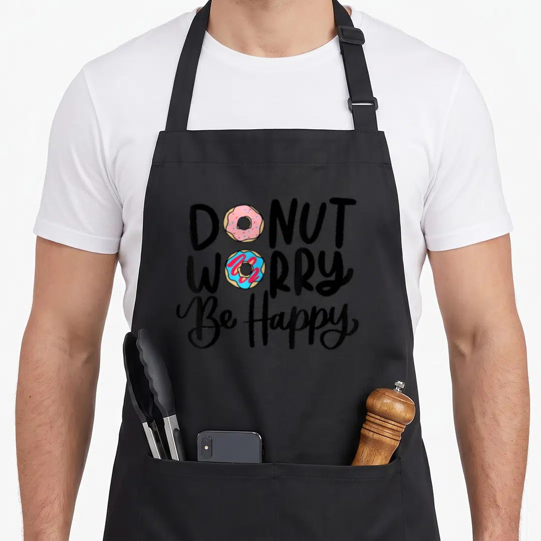 Candy donut apron - Two donuts replacing O's in DONUT WORRY Be Happy text — shown on a black cotton canvas apron against a white background