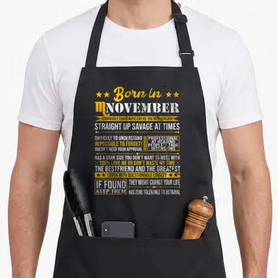 November scorpio birthday apron - Yellow and white text listing Scorpio traits like sarcasm and loyalty with stars — shown on a black cotton canvas apron against a white background