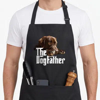 Dog chocolate brown apron - Lying brown dog with white The Dogfather text overlaid on chest — shown on a black cotton canvas apron against a white background