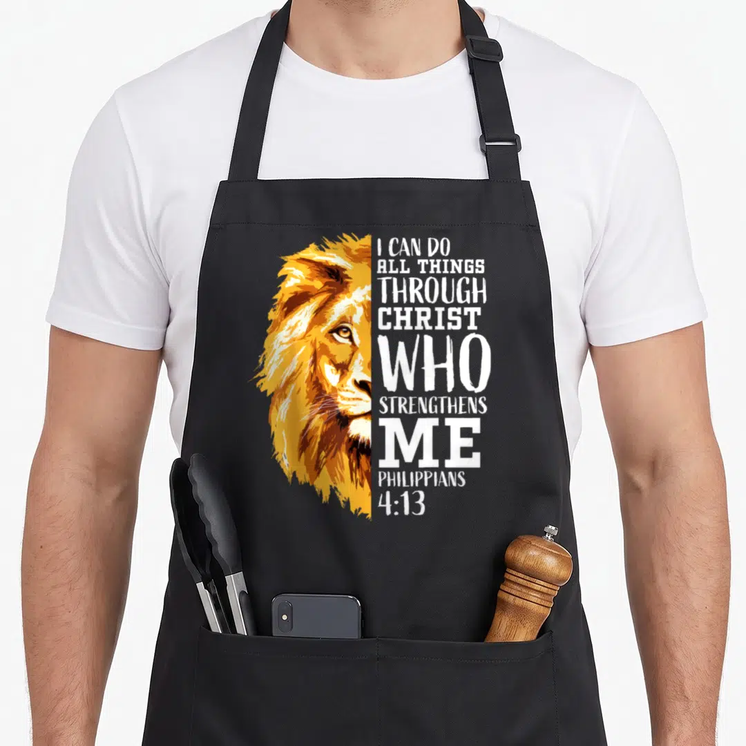 Lion bible verse apron - Realistic lion half-face next to stacked white Bible verse text in orange and yellow tones — shown on a black cotton canvas apron against a white background