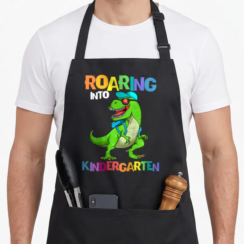 Kindergarten rex dinosaur apron - Green T-Rex with cap sunglasses backpack and multicolored ROARING INTO KINDERGARTEN — shown on a black cotton canvas apron against a white background