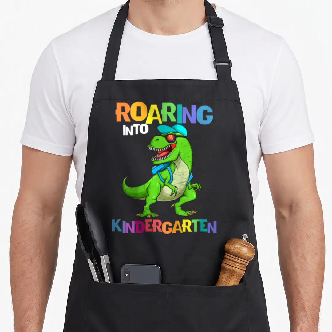 Kindergarten rex dinosaur apron - Green T-Rex with cap sunglasses backpack and multicolored ROARING INTO KINDERGARTEN — shown on a black cotton canvas apron against a white background