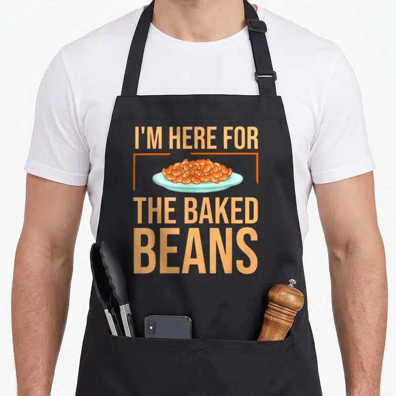 Canned baked beans apron - I'm Here For The Baked Beans text surrounding an orange bean pile on a light blue plate — shown on a black cotton canvas apron against a white background