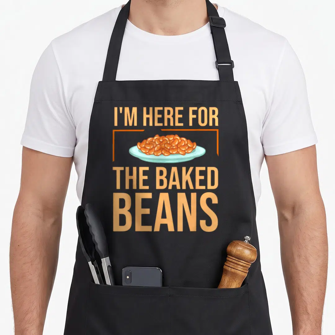 Canned baked beans apron - I'm Here For The Baked Beans text surrounding an orange bean pile on a light blue plate — shown on a black cotton canvas apron against a white background
