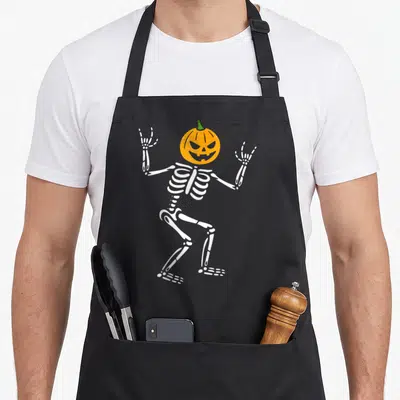 Skeleton pumpkin halloween apron - Cartoon white skeleton with orange pumpkin head making rock-on hand gestures — shown on a black cotton canvas apron against a white background