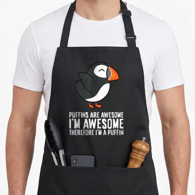 Puffin parrot v neck apron - Cartoon puffin in black white and orange above stacked white text reading PUFFINS ARE — shown on a black cotton canvas apron against a white background