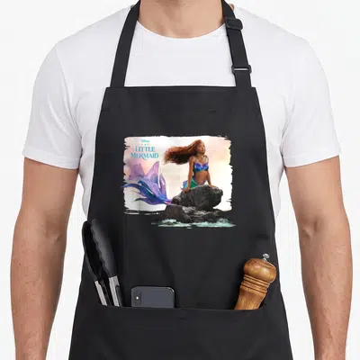 Live mermaid ariel apron - The Little Mermaid sitting on grey rock with purple blue tail and title text — shown on a black cotton canvas apron against a white background