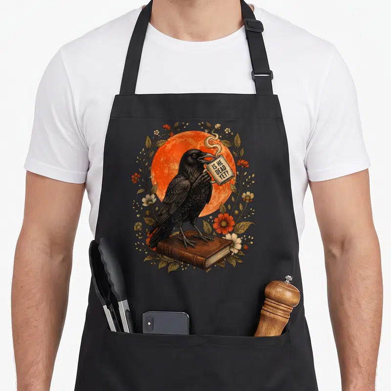 Dead gothic apron - Realistic crow on brown book holding mug with IS HE DEAD YET text framed by flowers and orange sun — shown on a black cotton canvas apron against a white background