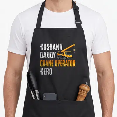Crane daddy apron - Stacked HUSBAND DADDY CRANE OPERATOR HERO text beside a yellow crane truck in distressed white — shown on a black cotton canvas apron against a white background