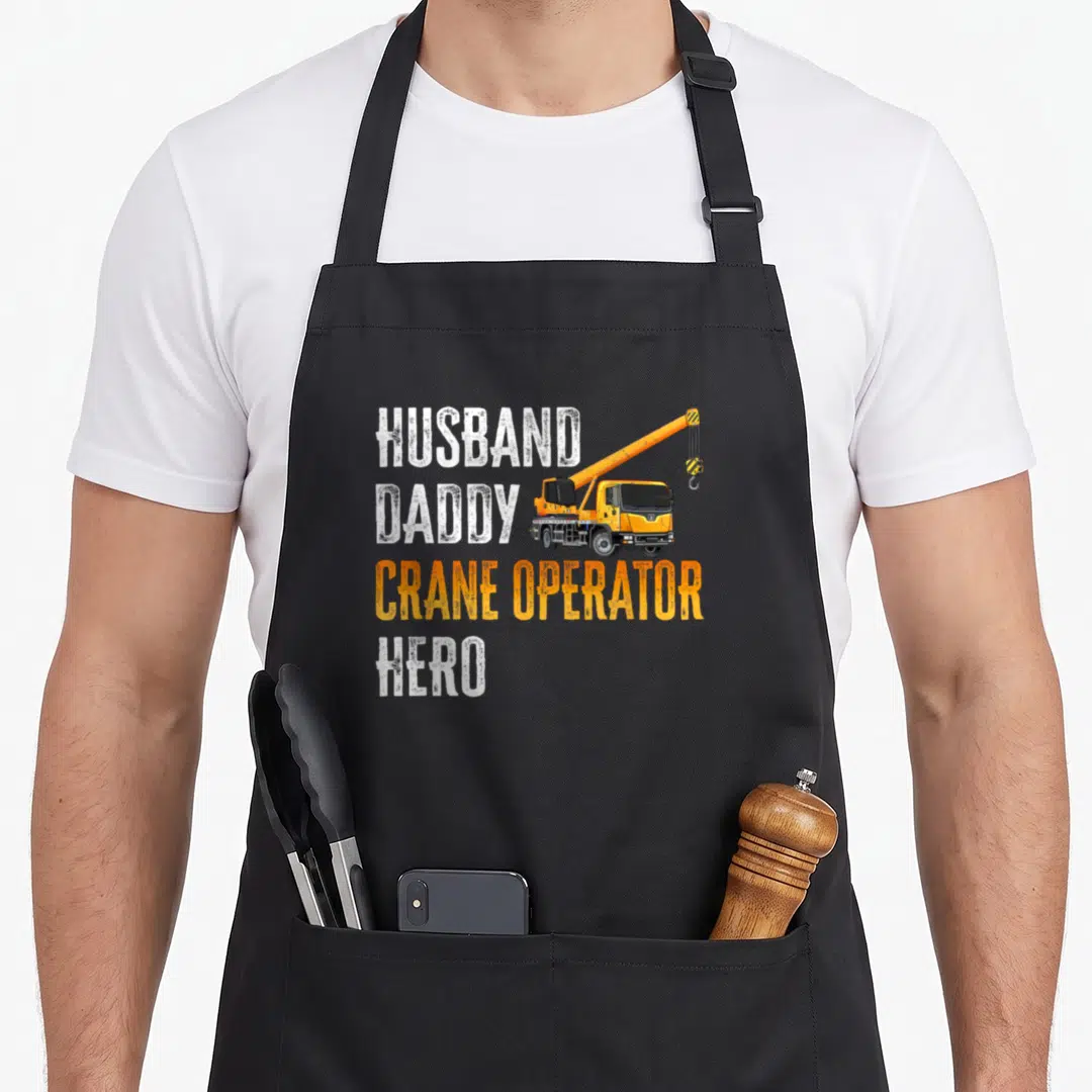 Crane daddy apron - Stacked HUSBAND DADDY CRANE OPERATOR HERO text beside a yellow crane truck in distressed white — shown on a black cotton canvas apron against a white background