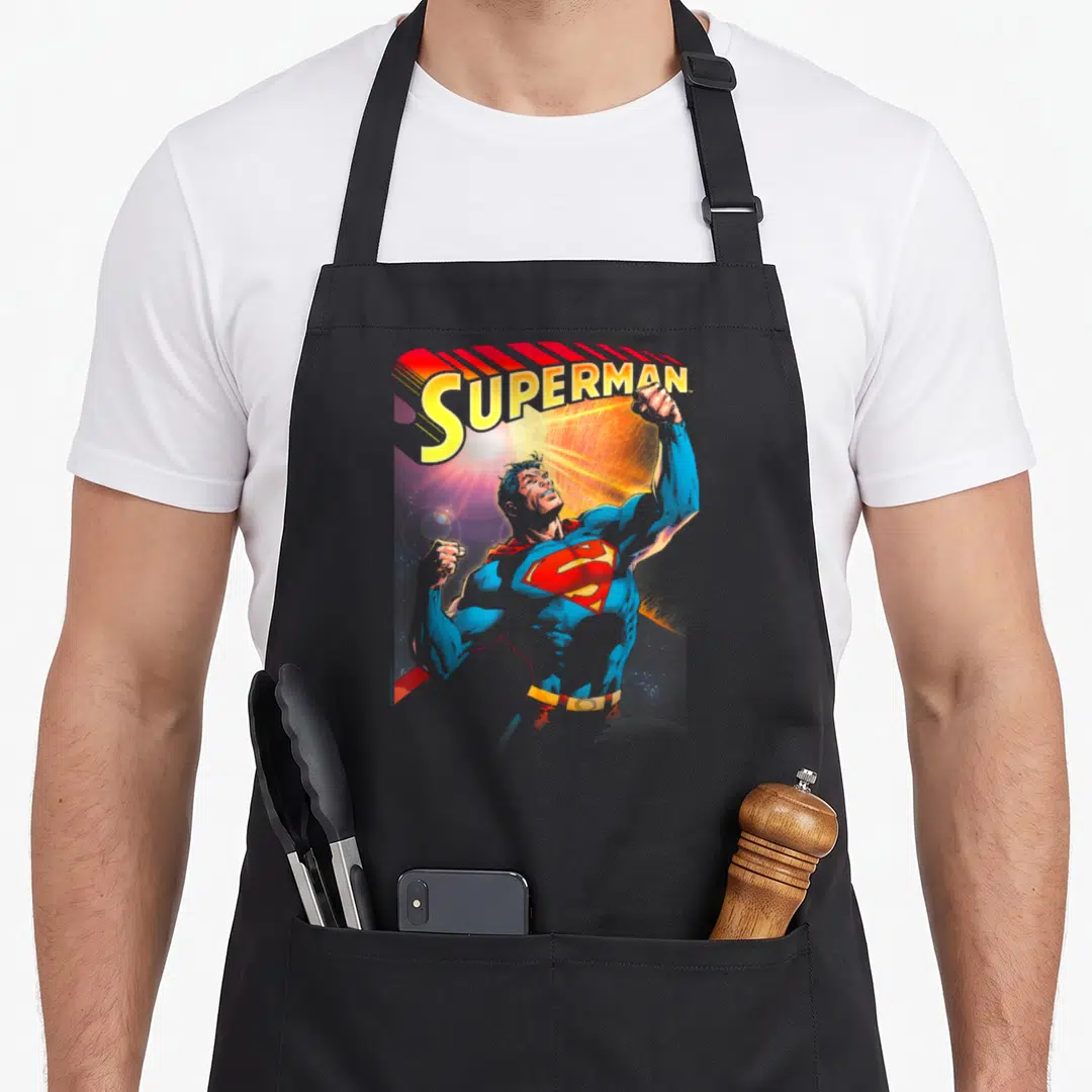 Trend apron for kids - Realistic Superman raising fists under yellow SUPERMAN text with red cape — shown on a black cotton canvas apron against a white background