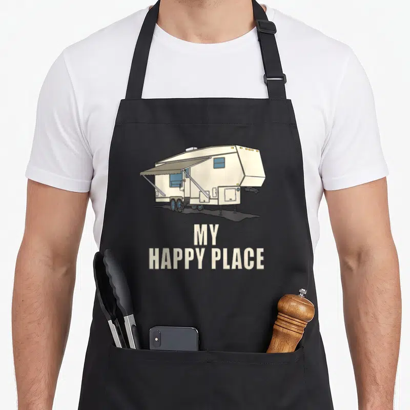 5th fifth wheel apron - Beige and blue cartoon RV with MY HAPPY PLACE text stacked below on grey background — shown on a black cotton canvas apron against a white background