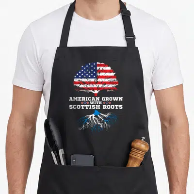 Root scotland apron - Tree with US flag leaves and Scottish flag roots text American Grown With Scottish Roots — shown on a black cotton canvas apron against a white background