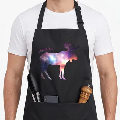 Alaska sky apron - Moose silhouette filled with purple blue orange pink galaxy colors standing over water reflection — shown on a black cotton canvas apron against a white background