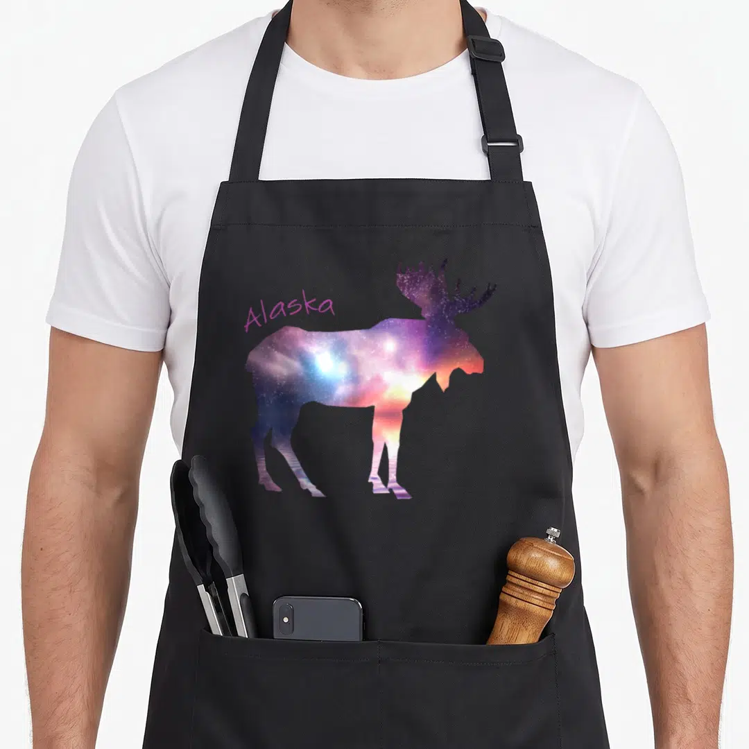 Alaska sky apron - Moose silhouette filled with purple blue orange pink galaxy colors standing over water reflection — shown on a black cotton canvas apron against a white background